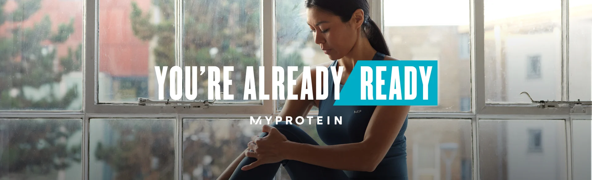 A woman wearing MP gym clothing sat on a window ledge stretching with the text 'You're Already Ready - MYPROTEIN' overlaying the image.